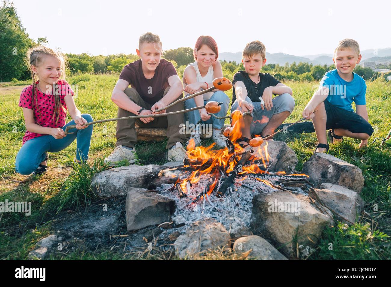 Group of Kids - Boys and girls cheerfully smiling and roasting sausages on long sticks over a campfire flame. Outdoor active time spending or camping Stock Photo