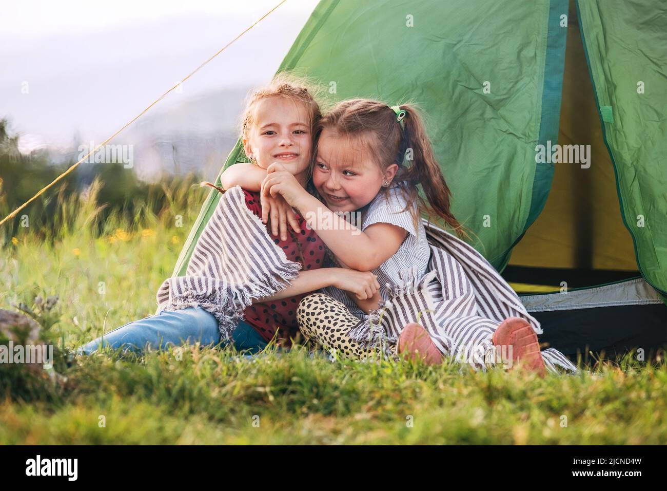 Two little hugging girls sisters sitting on the green grass next camp tent entrance, cheerfully smiling. Careless childhood, family values and outdoor Stock Photo