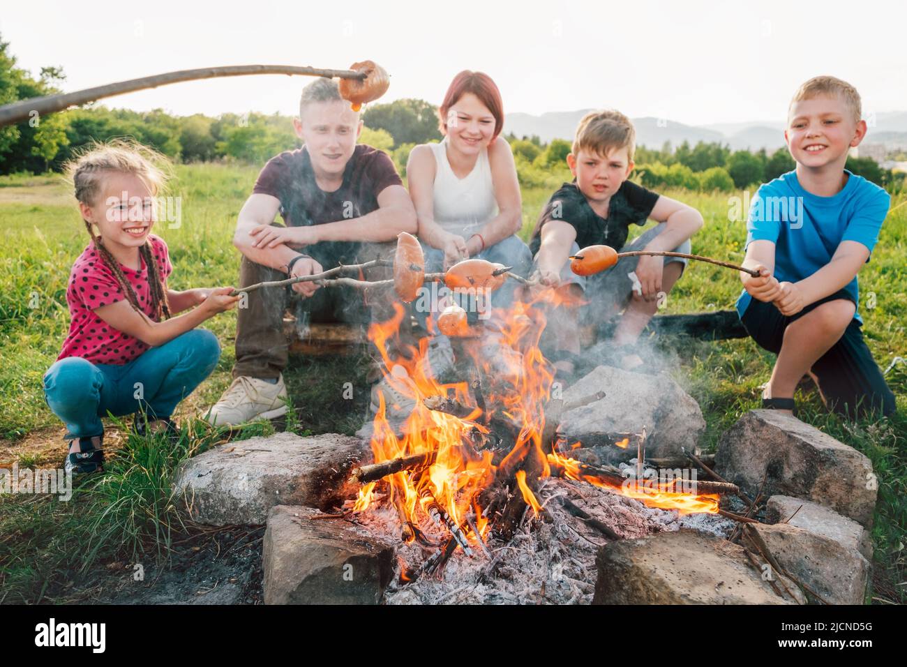 Group of Kids - Boys and girls cheerfully smiling and roasting sausages on sticks over a campfire flame Sausages selective focus. Outdoor active time Stock Photo