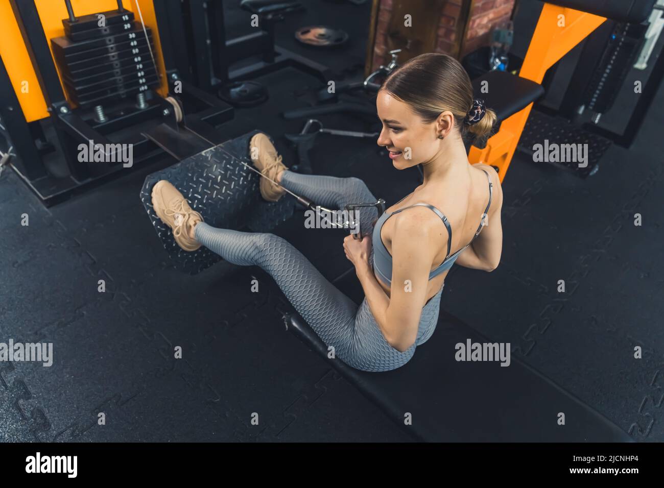 Active strong European millennial woman in gray fashionable sportswear exercising on a leg muscles machine. High quality photo Stock Photo