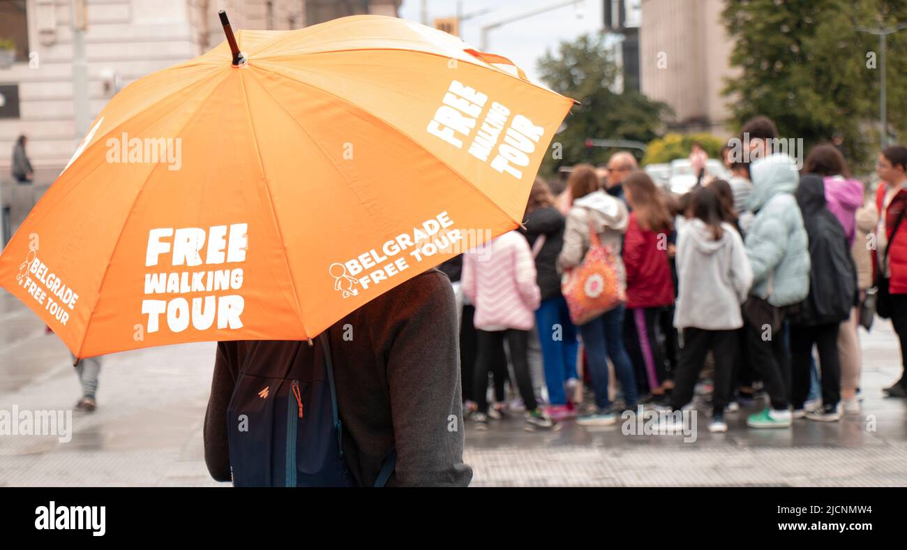 Belgrade, Serbia - May 28, 2022: Tour guide standing under orange umbrella with Free walking tour sign, on a city square on a rainy day, rear view and Stock Photo
