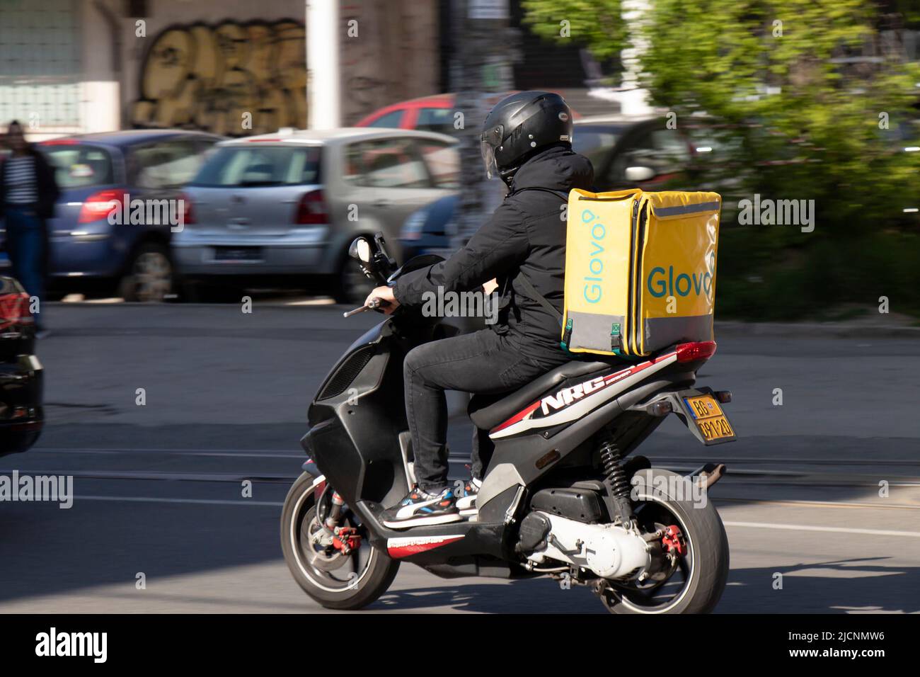 Belgrade, Serbia - April, 15, 2022: Courier working for Glovo food delivery service riding  scooter with container on his back, on city street Stock Photo