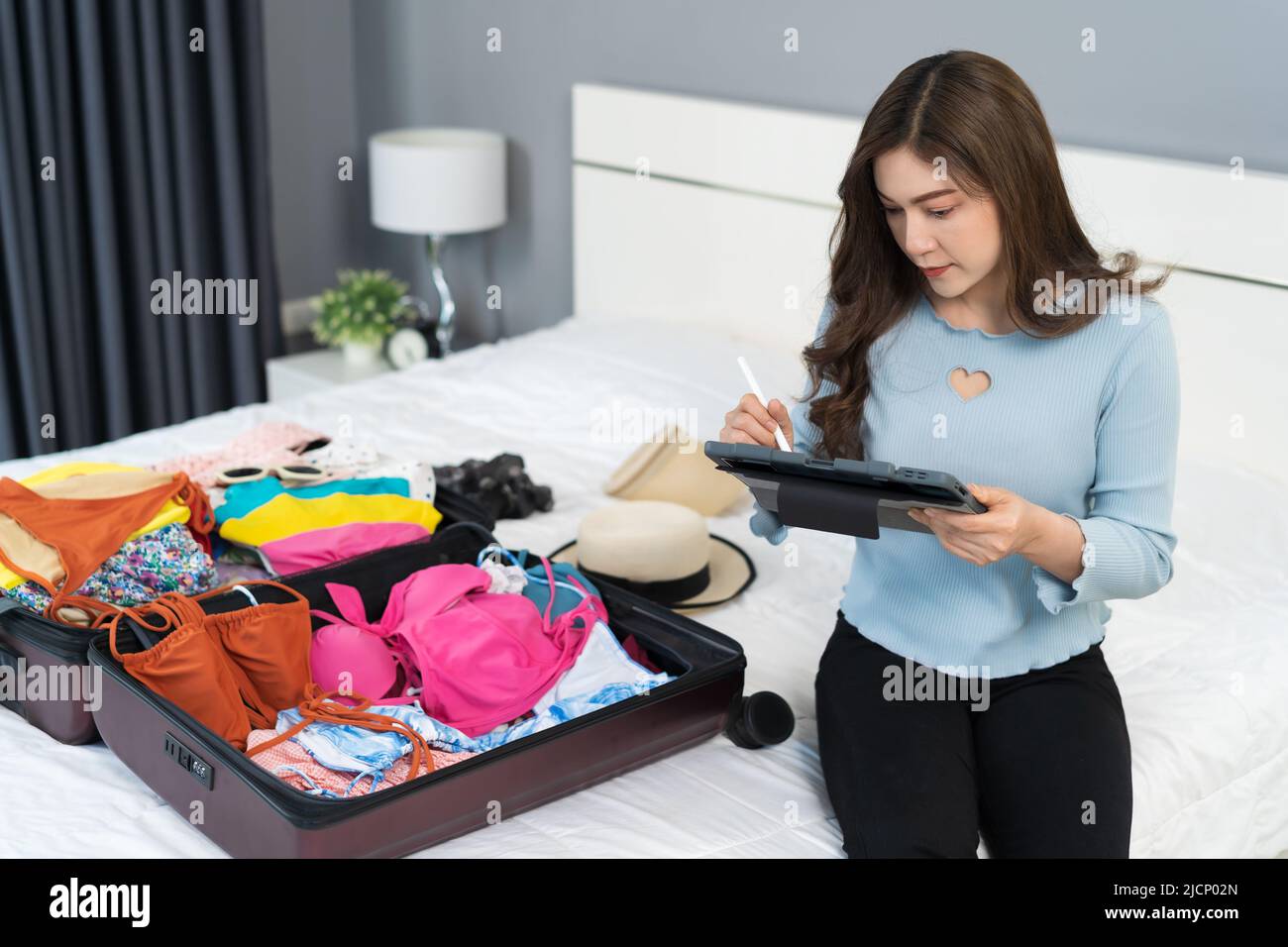 young woman writing tablet and packing clothes into suitcase on a bed at home, planning travel holiday. Stock Photo