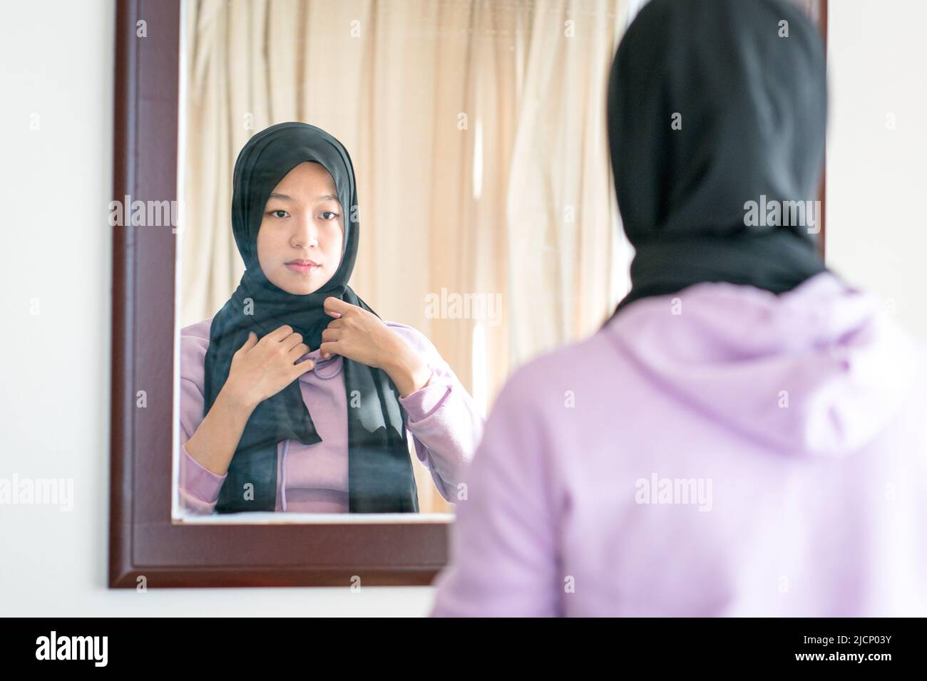 Reflection on a wall mirror of a muslim woman wearing and adjusting her head scarf. Stock Photo