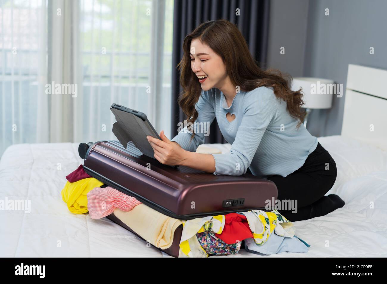 cheerful woman using tablet and packing clothes into suitcase on a bed at home, planning travel holiday. Stock Photo