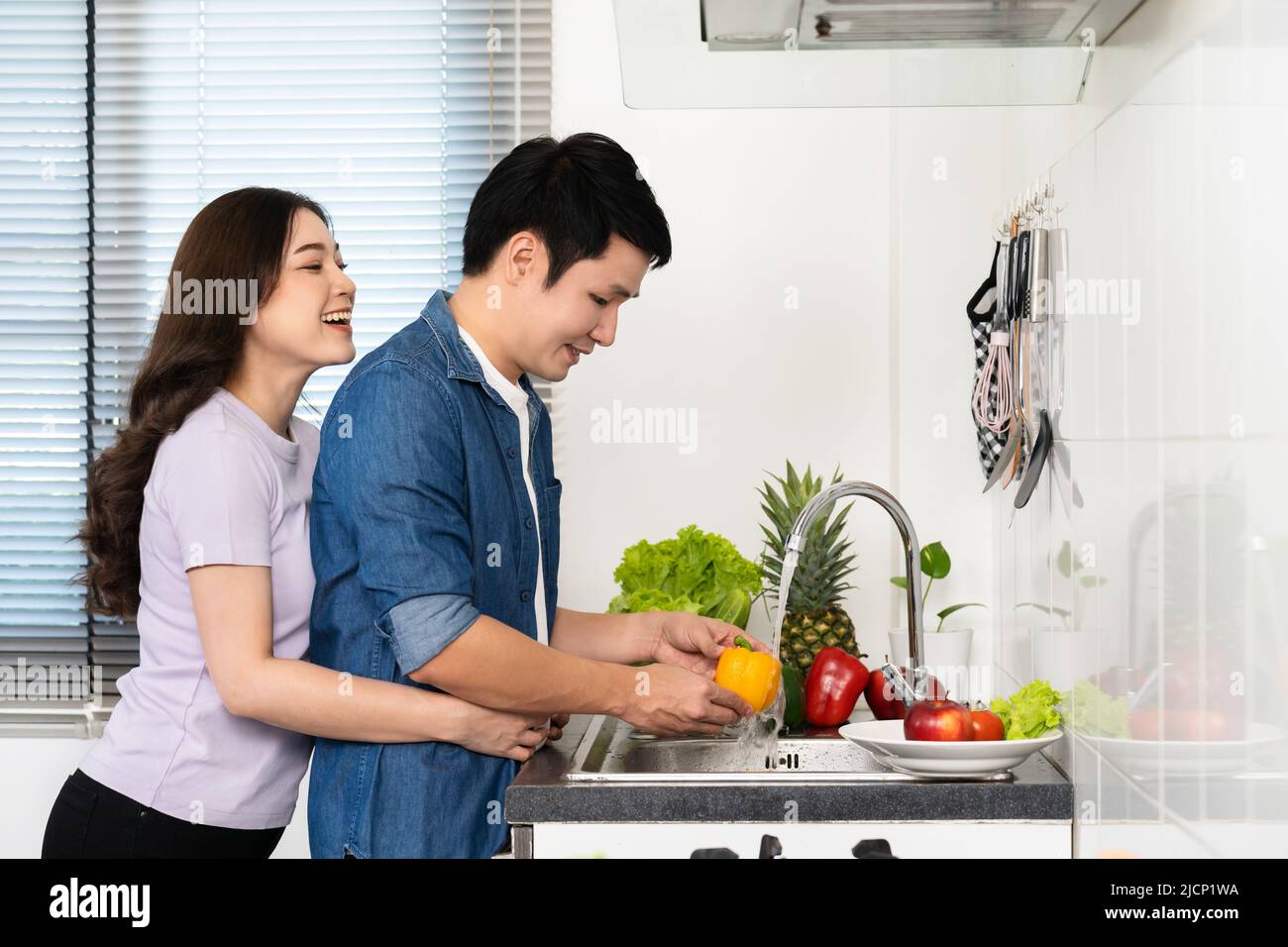 happy young couple washing sweet pepper and vegetables in the sink in the kitchen at home. woman is hugging man Stock Photo