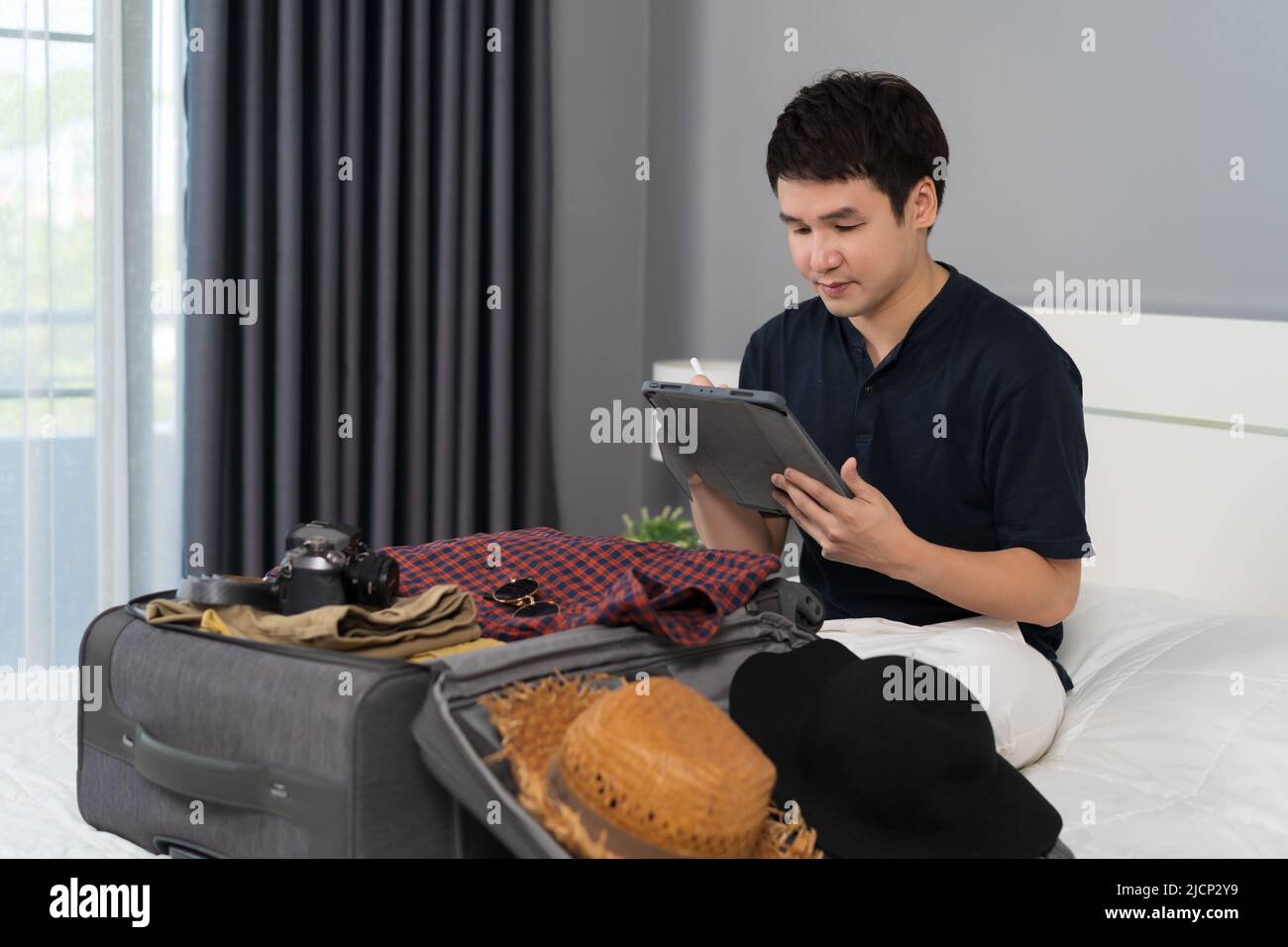 young man writing tablet and packing clothes into suitcase on a bed at home, planning travel holiday. Stock Photo