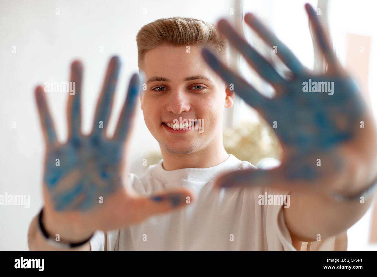 Beautiful blonde toothy smile young man drawing. Male looking at camera and show defocused blue painted palms. Stock Photo