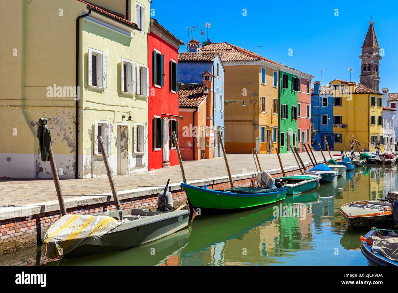 View of boats on narrow canal and colorful houses under blue sky in Burano, Italy. Stock Photo