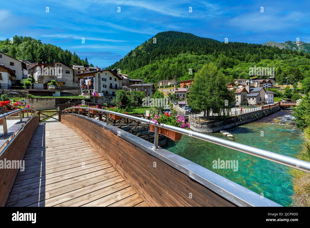 Narrow wooden bridge over alpine creek as mountains under blue sky on background in town of Pietraporzio in Piedmont, Northern Italy. Stock Photo