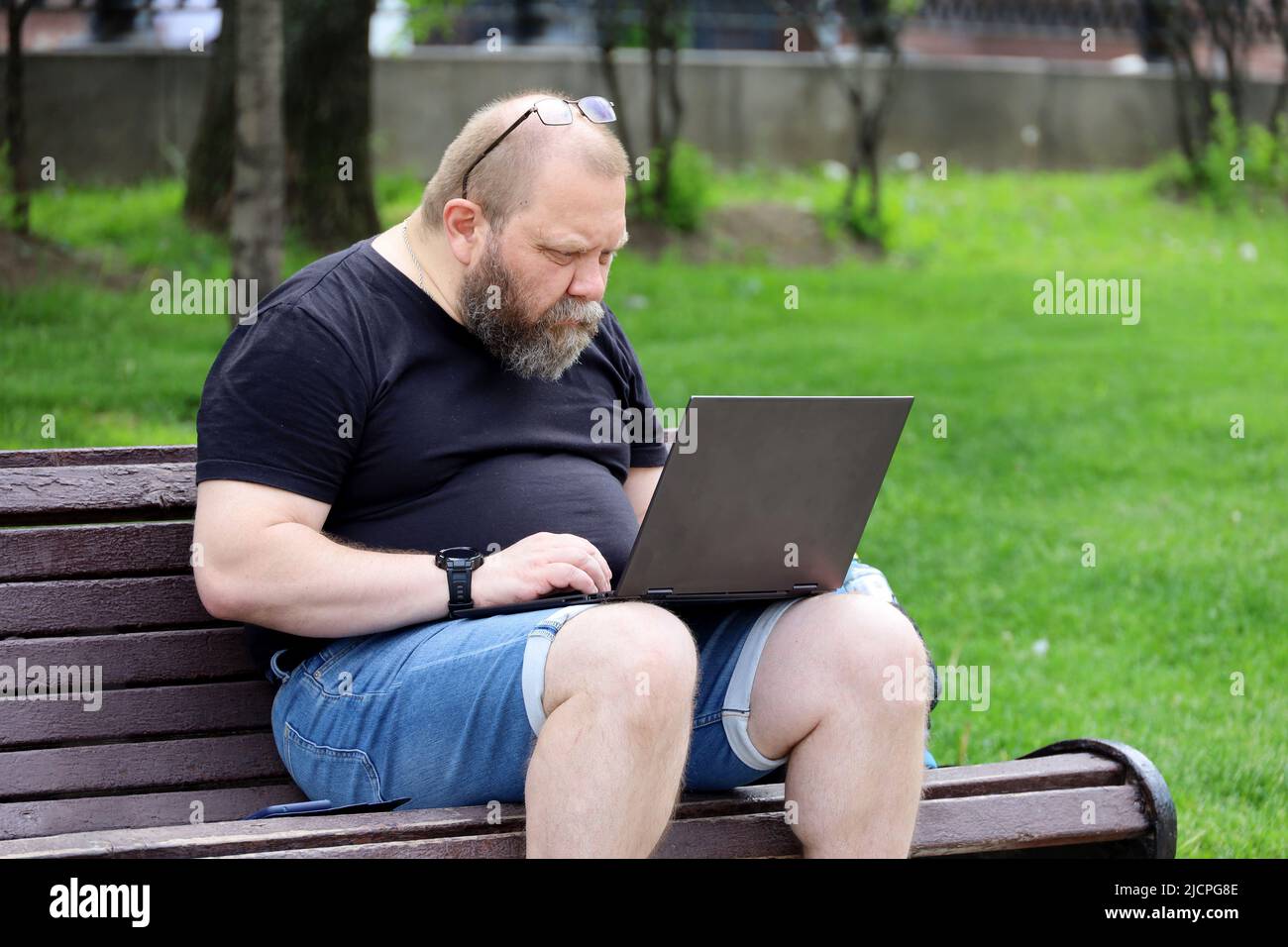 Bearded man in jeans shorts sitting with a laptop on a bench in summer park. Concept of freelancer working outdoors Stock Photo