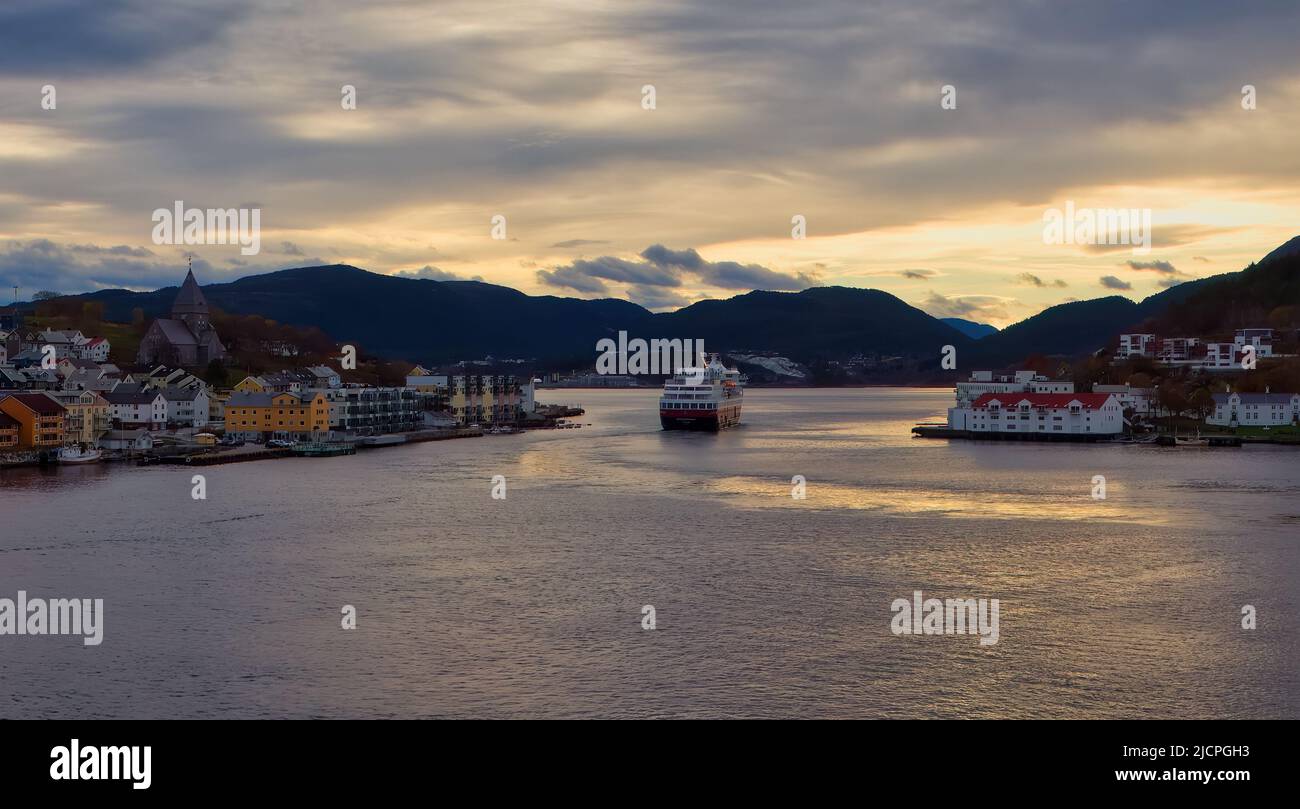 A Hurtigruten cruise ship heads out of the port of Kristiansund into the early morning light as day breaks over the Norwegain fjords. Stock Photo