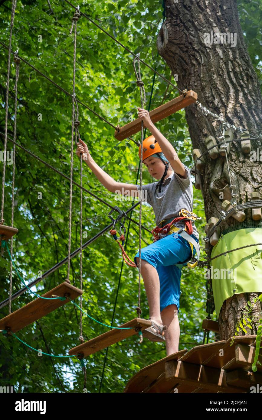 Rope park. A boy teenager in a helmet walks on suspended rope ladders. Carabiners and safety straps. Safety. Summer activity. Sport. Children's playground in nature in the forest. Stock Photo