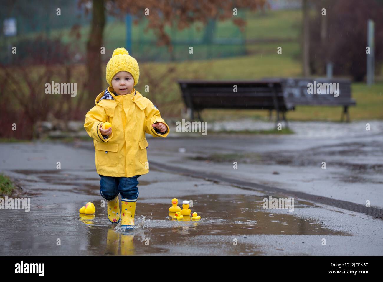 Beautiful funny blonde toddler boy with rubber ducks and colorful umbrella, jumping in puddles and playing in the rain, wintertime Stock Photo