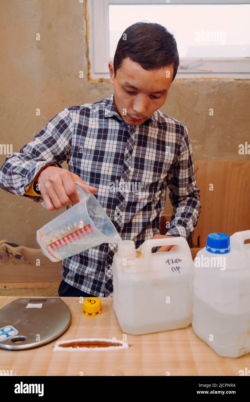 Carpenter pouring epoxy resin into liquid cup. Process of making a craft resin and wood table