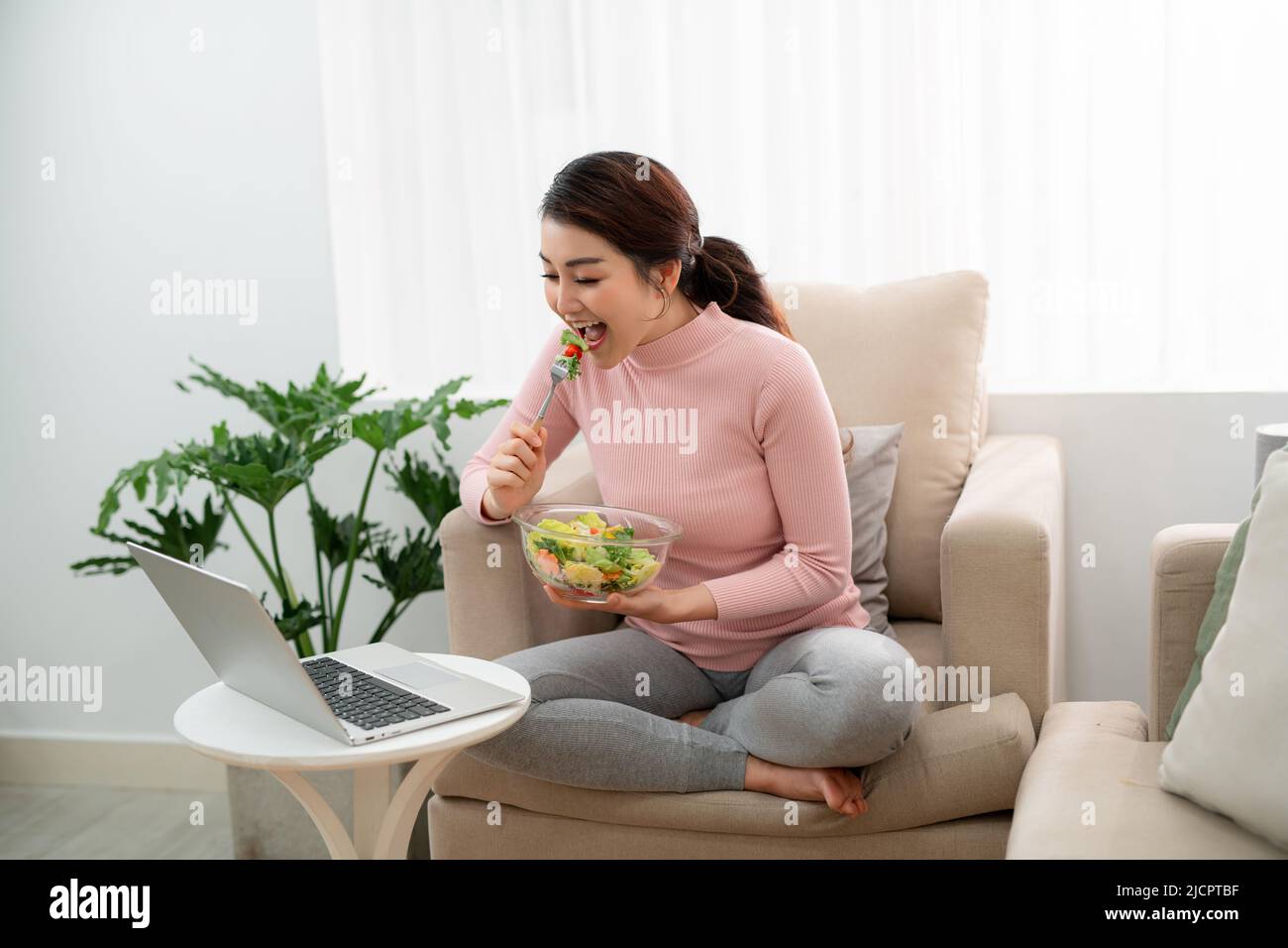 Beautiful young woman eating a healthy salad in living room, using a ...