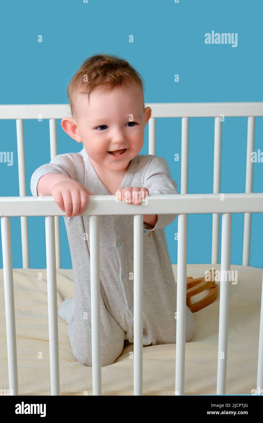 Happy infant baby boy stands in the crib, studio blue background