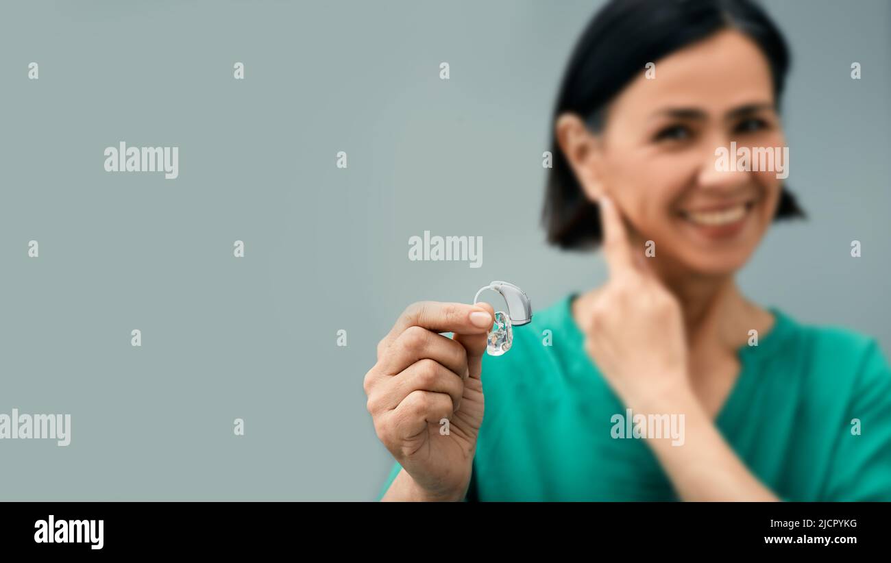 Mature woman holding BTE hearing aid in hand on foreground and points to her ear. Treatment of deafness in adult people Stock Photo