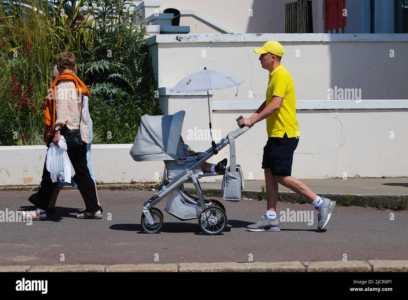 Hastings, East Sussex, UK. 15 Jun, 2022. UK Weather: Hot and sunny at the seaside town of Hastings in East Sussex as Brits enjoy the warm weather today along the seafront promenade. Man in a bright yellow top and cap pushing a stroller in the hot and sunny weather. Photo Credit: Paul Lawrenson /Alamy Live News Stock Photo