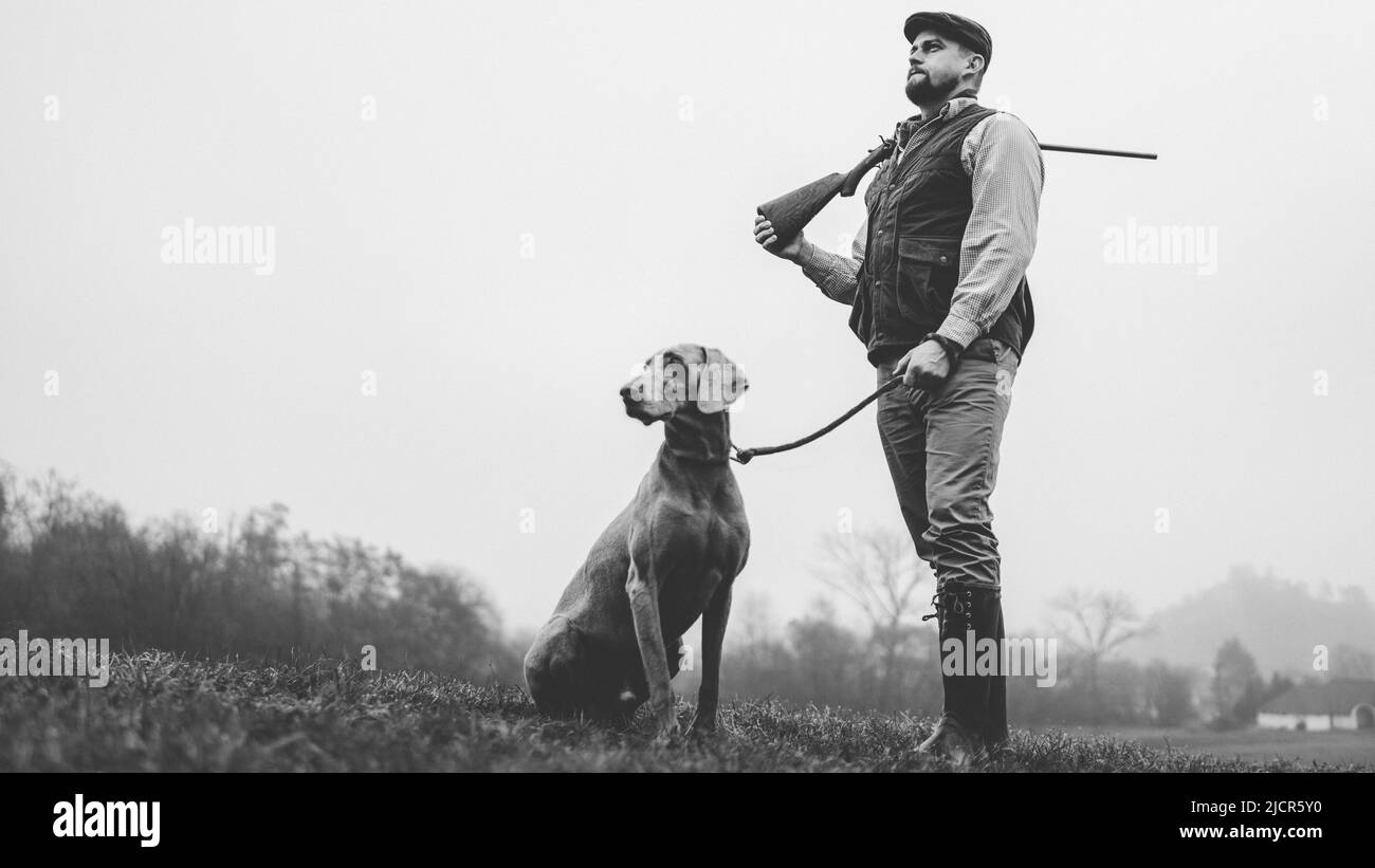Hunter man with dog in traditional shooting clothes on field holding shotgun, black and white photo. Stock Photo