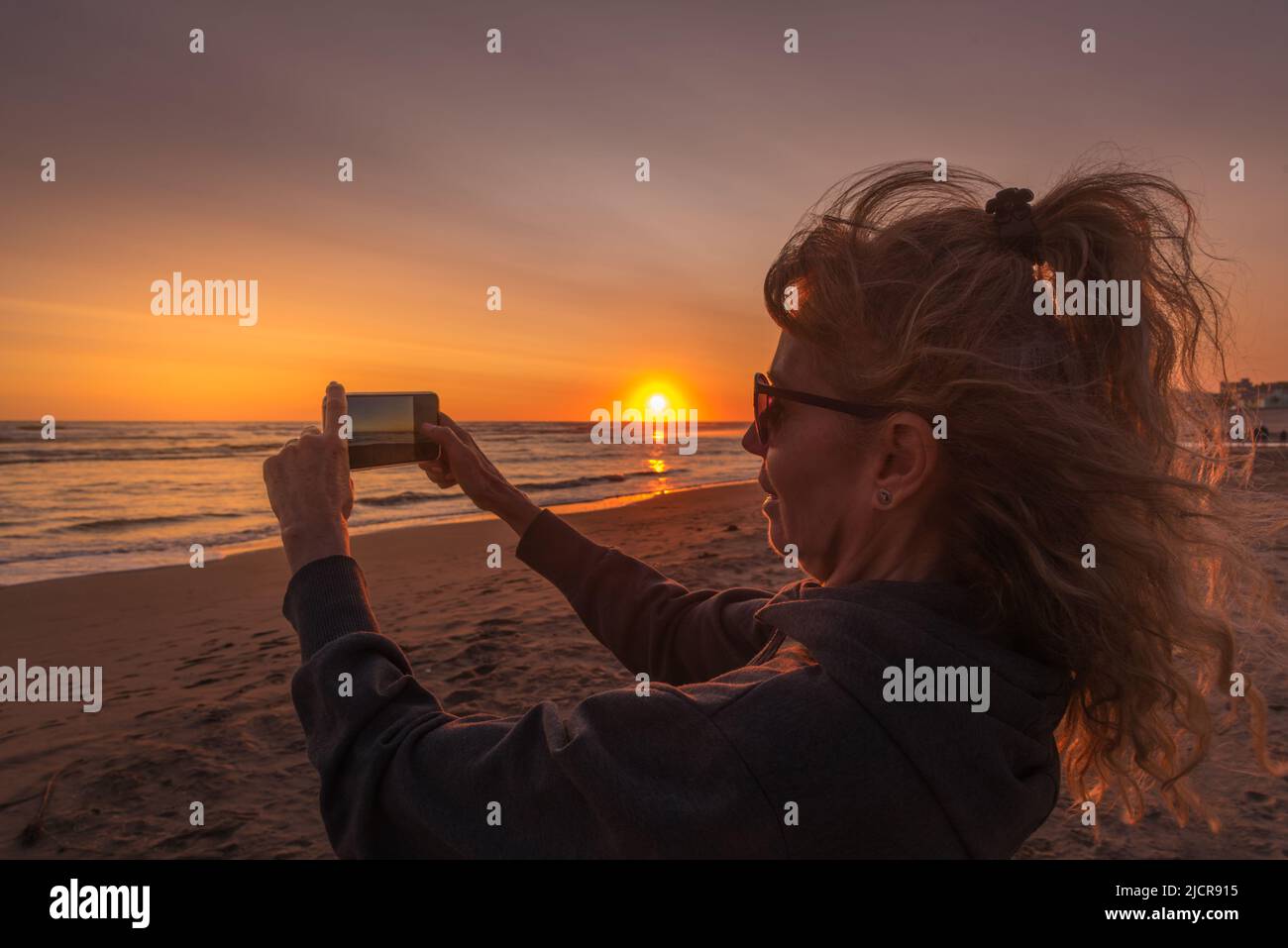 Mature woman taking a photo with her smartphone at sunset with the sun over the sea. Stock Photo