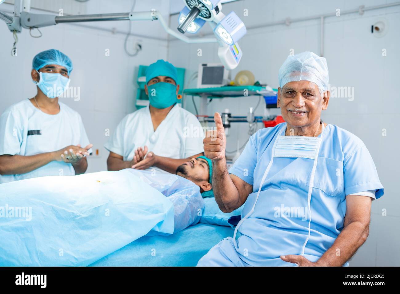 Senior surgeon showing thumbs up while practitioners applauding from behind at operating theatre - concept of successful treatment, encouraging and Stock Photo