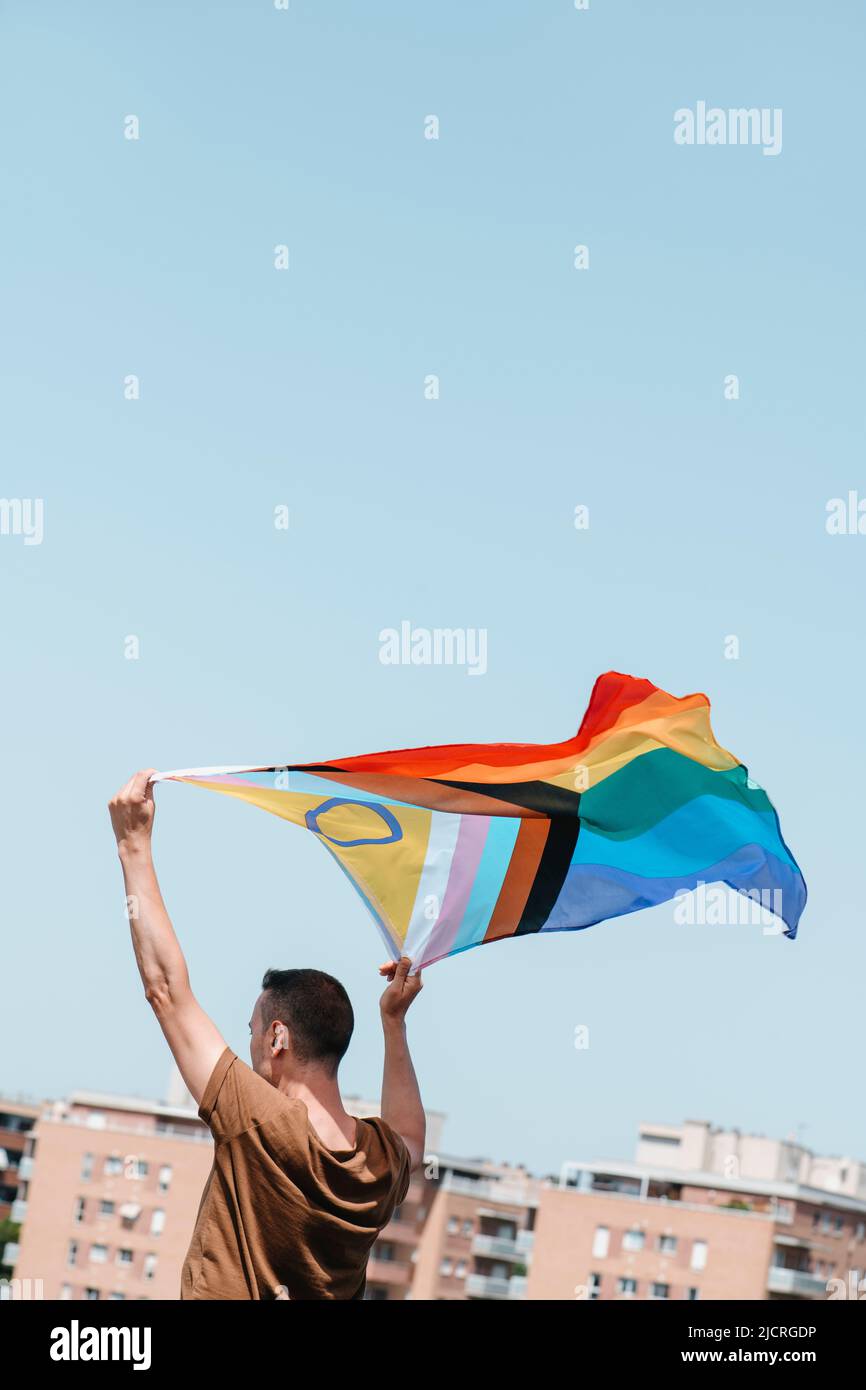 a young man waves an intersex-inclusive progress pride flag above his head on the blue sky, in the city Stock Photo