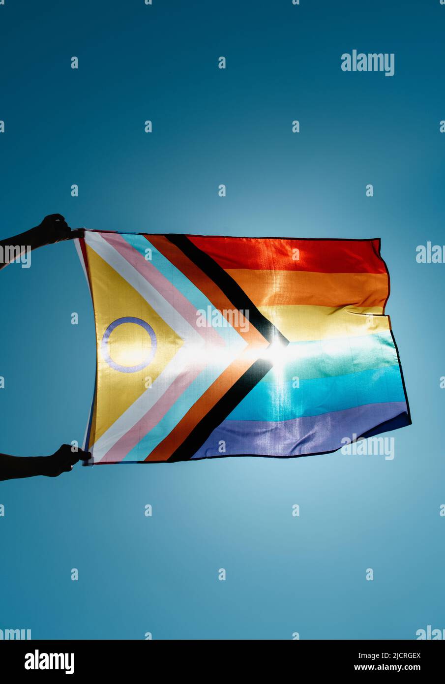 closeup of a man waving an intersex-inclusive progress pride flag in the air on the blue sky, with the sun in the background Stock Photo