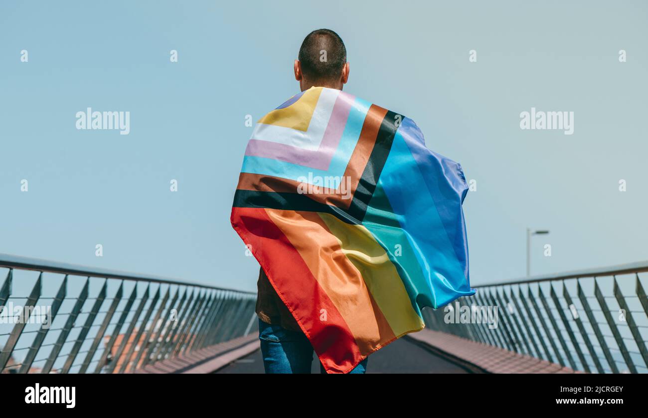 closeup of a man, seen from behind, wrapped in an intersex-inclusive progress pride flag walking by a bridge Stock Photo