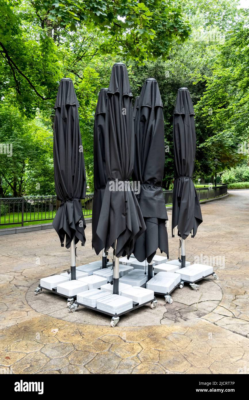black closed patio umbrella Black closed umbrellas of an empty cafe in green summer park. Outdoor  exterior furniture. Vertical view Stock Photo