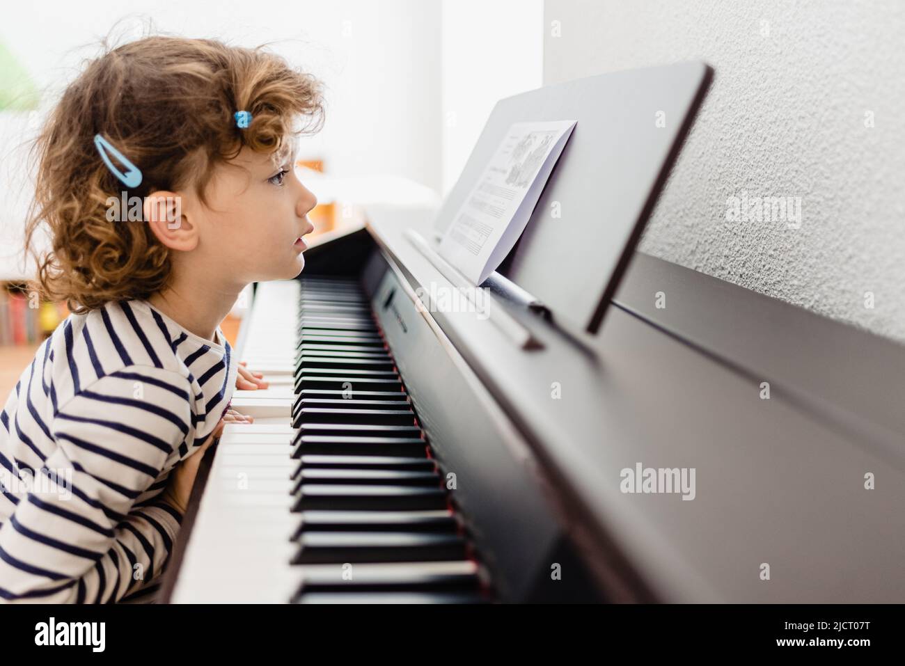 Pretty 3-year-old girl plays the keys of a piano and reads the sheet music with difficulty. Stock Photo