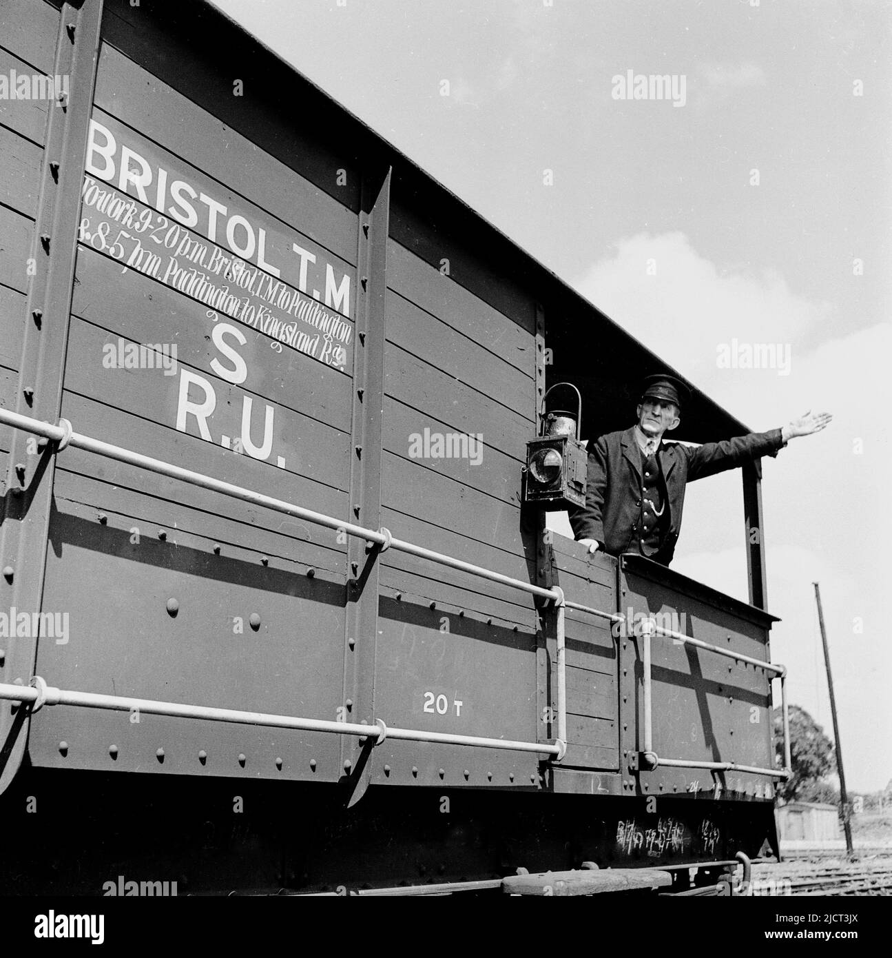 1950s, historical, a uniformed train guard in the open rear of a freight wagon, standing with his arm out, signalling, London docks, England UK. Writing on the side of the freight carriage, Bristol T. M, (Temple Mead) -Paddington. A railway warning lantern is hanging on the side of the wooden carriage. Stock Photo