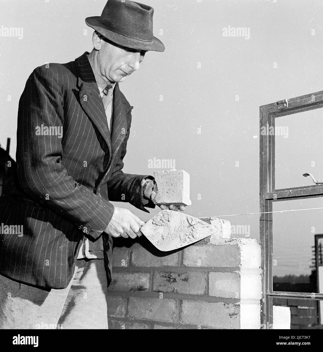 1950s, historical, elderly bricklayer wearing a shirt & tie, a pin-striped jacket and hat, trowel in hand, building a wall, England, UK. Stock Photo