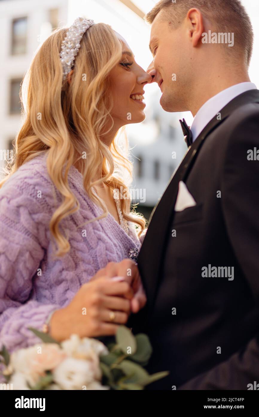 Beautiful young wedding couple in elegant outfits walk around city, smile and kiss on street background. Portrait side view closeup of happy bride and Stock Photo