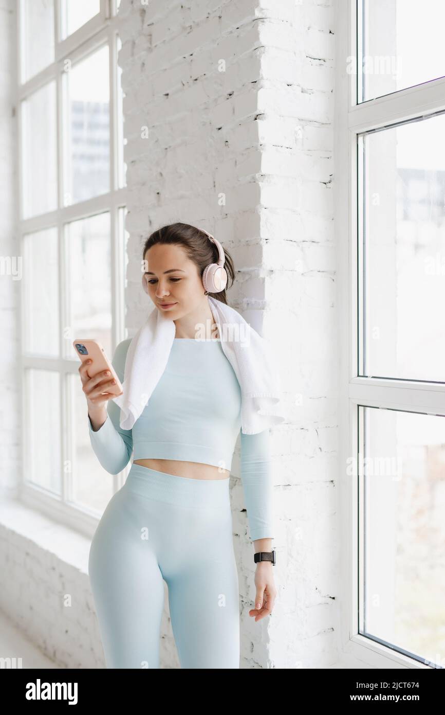 Sporty young woman in blue sportswear using smartphone and headphone resting after exercising leaning against a wall. Stock Photo