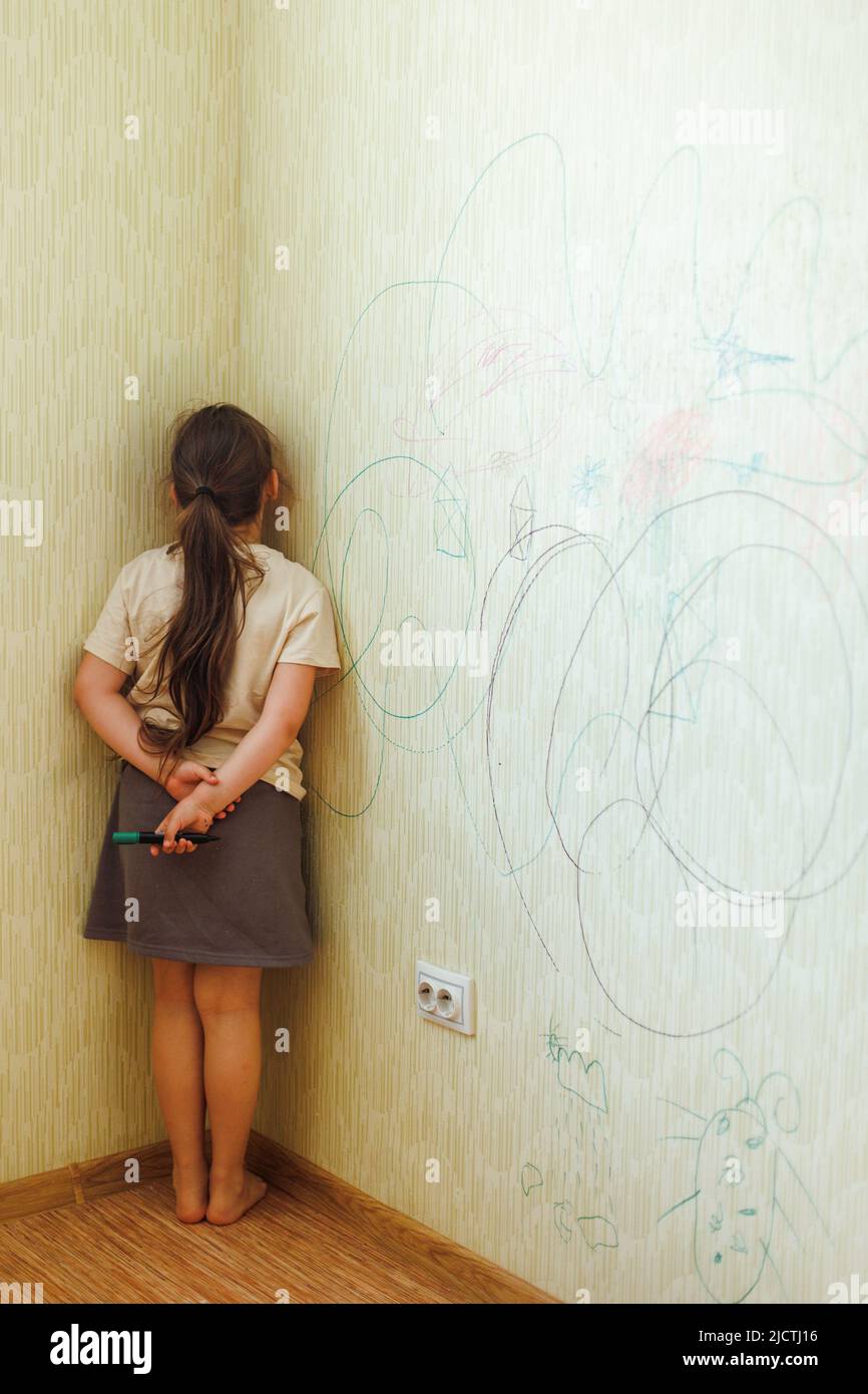 Portrait of little naughty girl holding marker behind back, standing punished in corner for spoilt wallpaper. Vertical. Stock Photo
