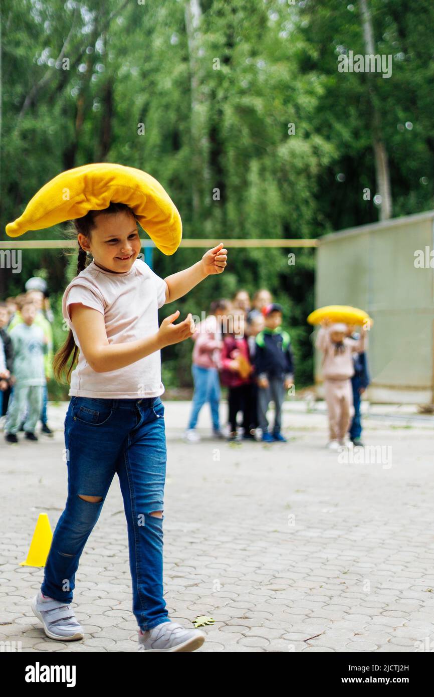 Smiling little girl carrying on yellow pillow in playground in green park. Summer holidays in camp, tourist center. Walking and playing outdoors Stock Photo