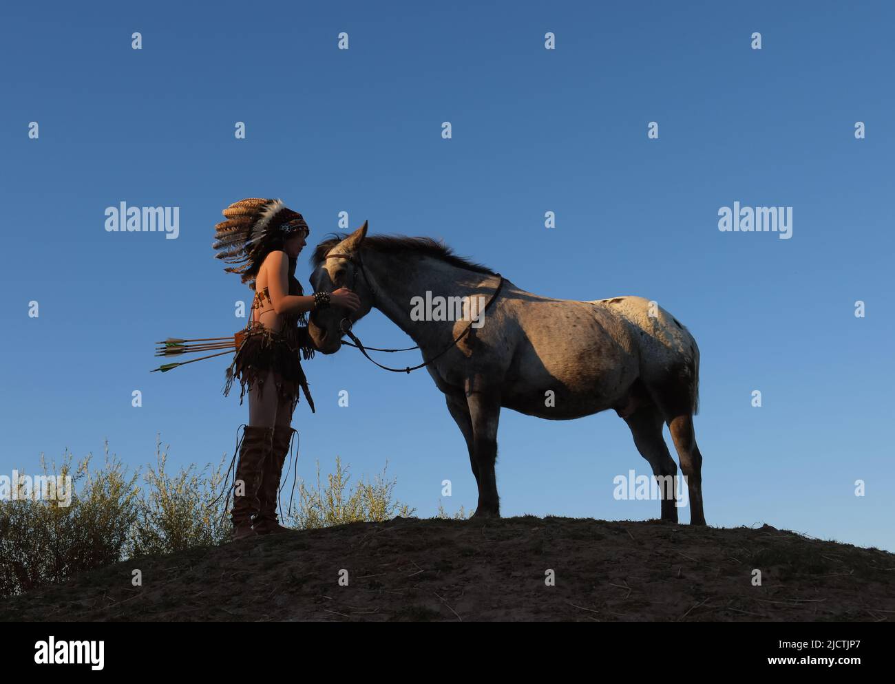 A young girl is seen at sunset as a Native American Indian.  She pauses on top of a small hill to cherish her horse Stock Photo