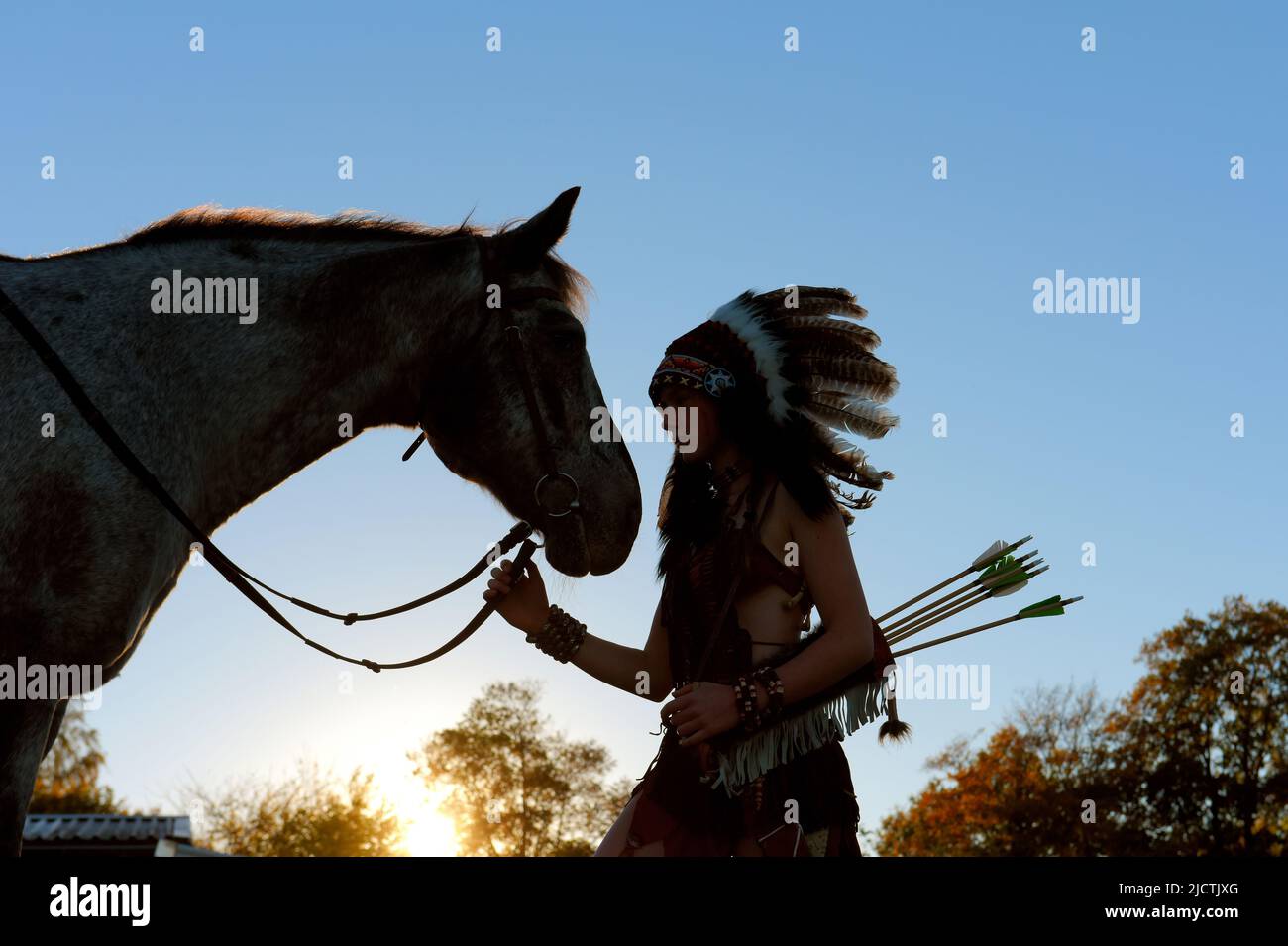 A young girl is seen at sunset as a Native American Indian.  She is seen together with her horse as the sun sets in the distance. Stock Photo