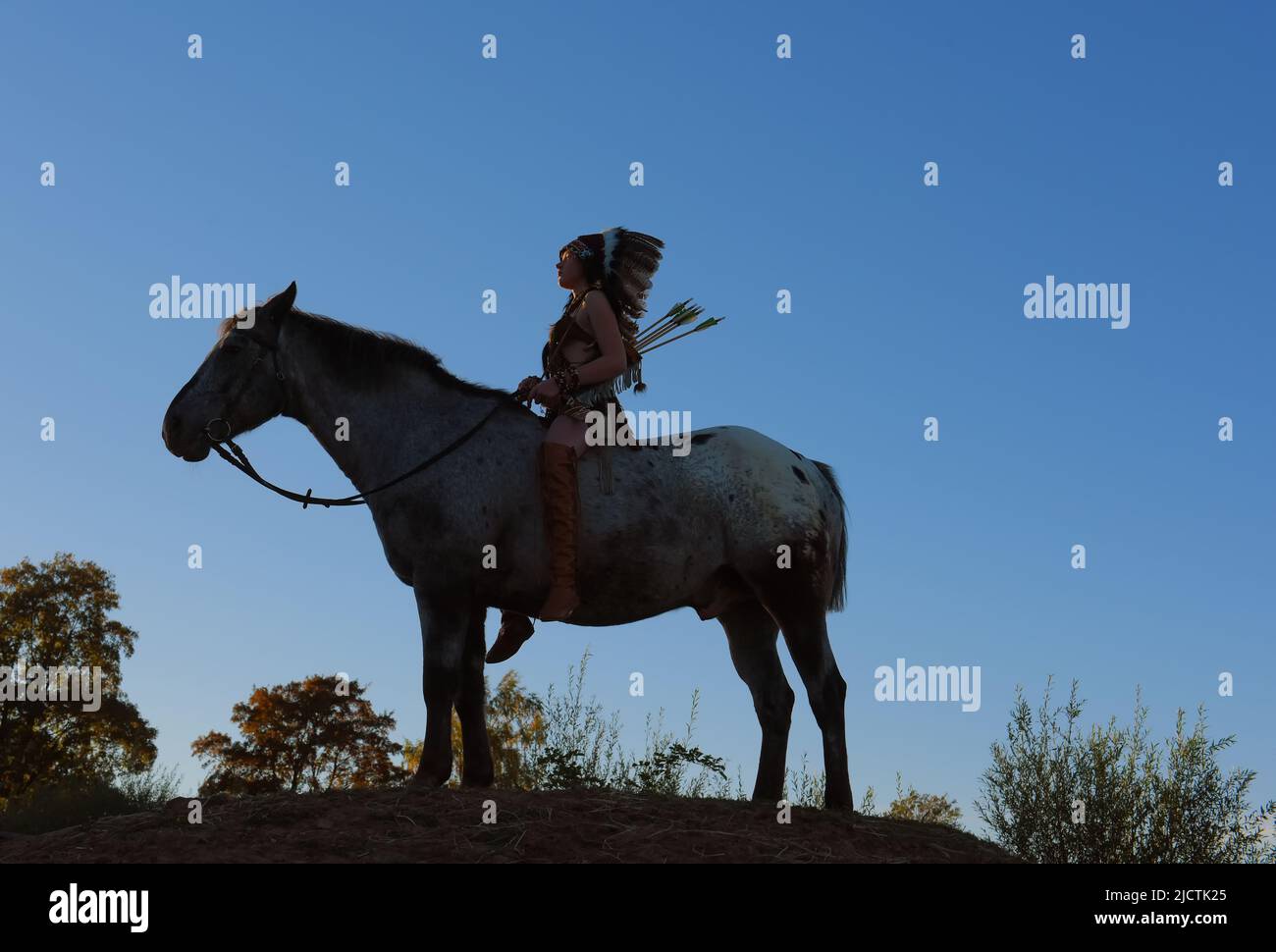 A young girl is seen at sunset as a Native American Indian.  She is seen together with her horse as the sun sets in the distance. Stock Photo