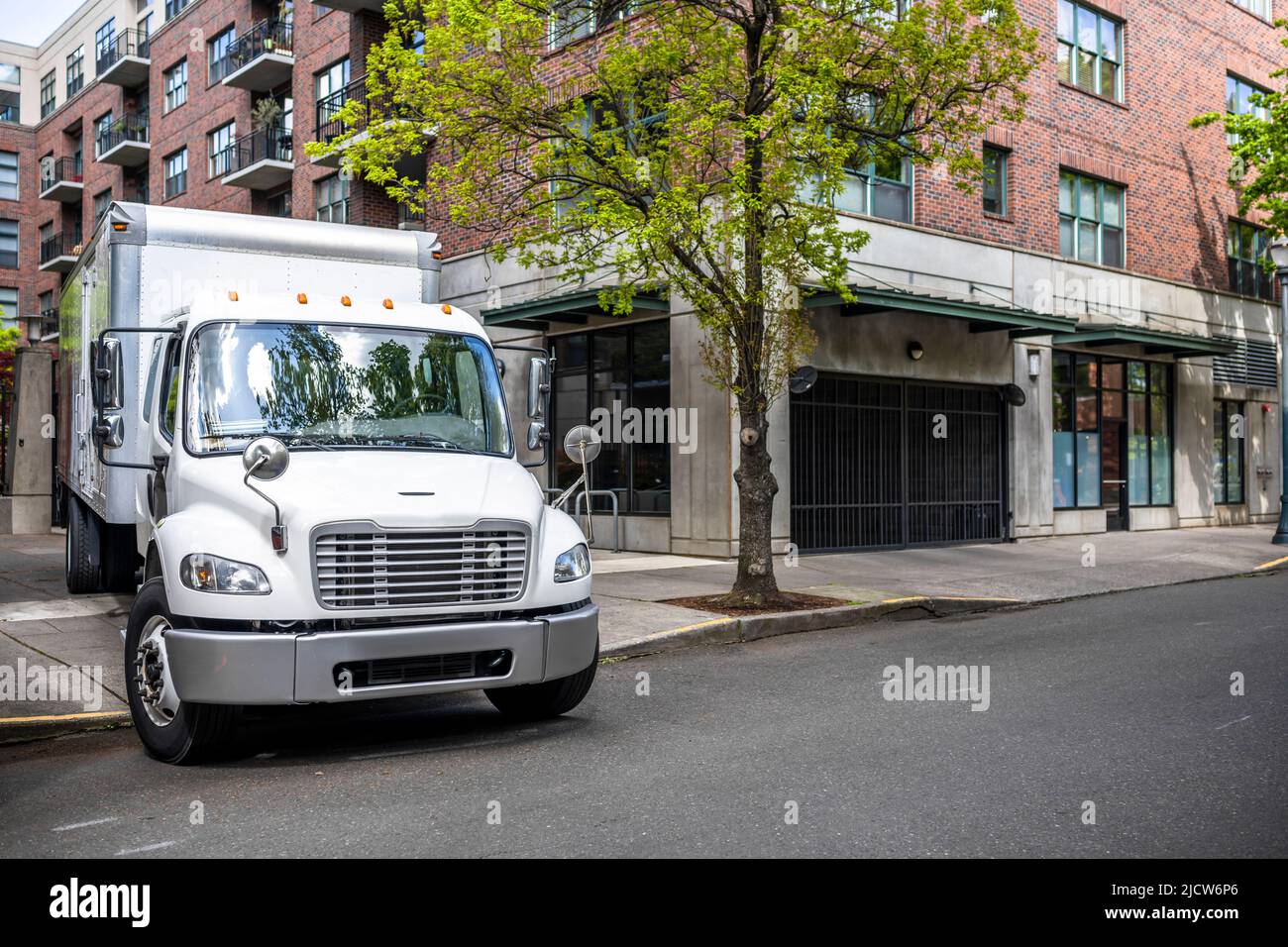 Big rig day cab white semi truck with long box trailer making local commercial delivery at urban city with multilevel residential apartments buildings Stock Photo