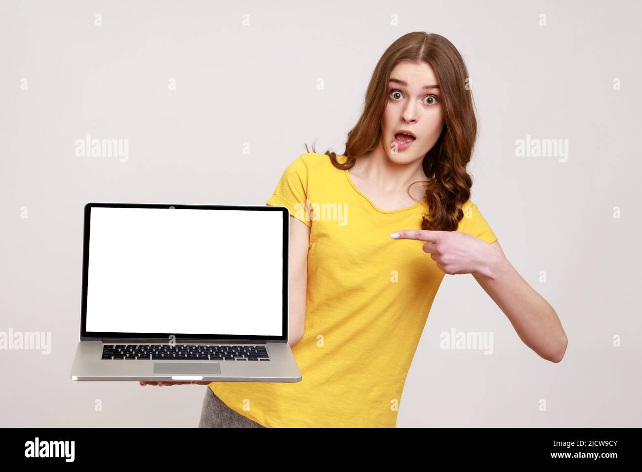 Shocking advertising. Portrait of amazed teenager girl in casual yellow T-shirt pointing at laptop with empty screen and looking surprised at camera. Indoor studio shot isolated on gray background. Stock Photo