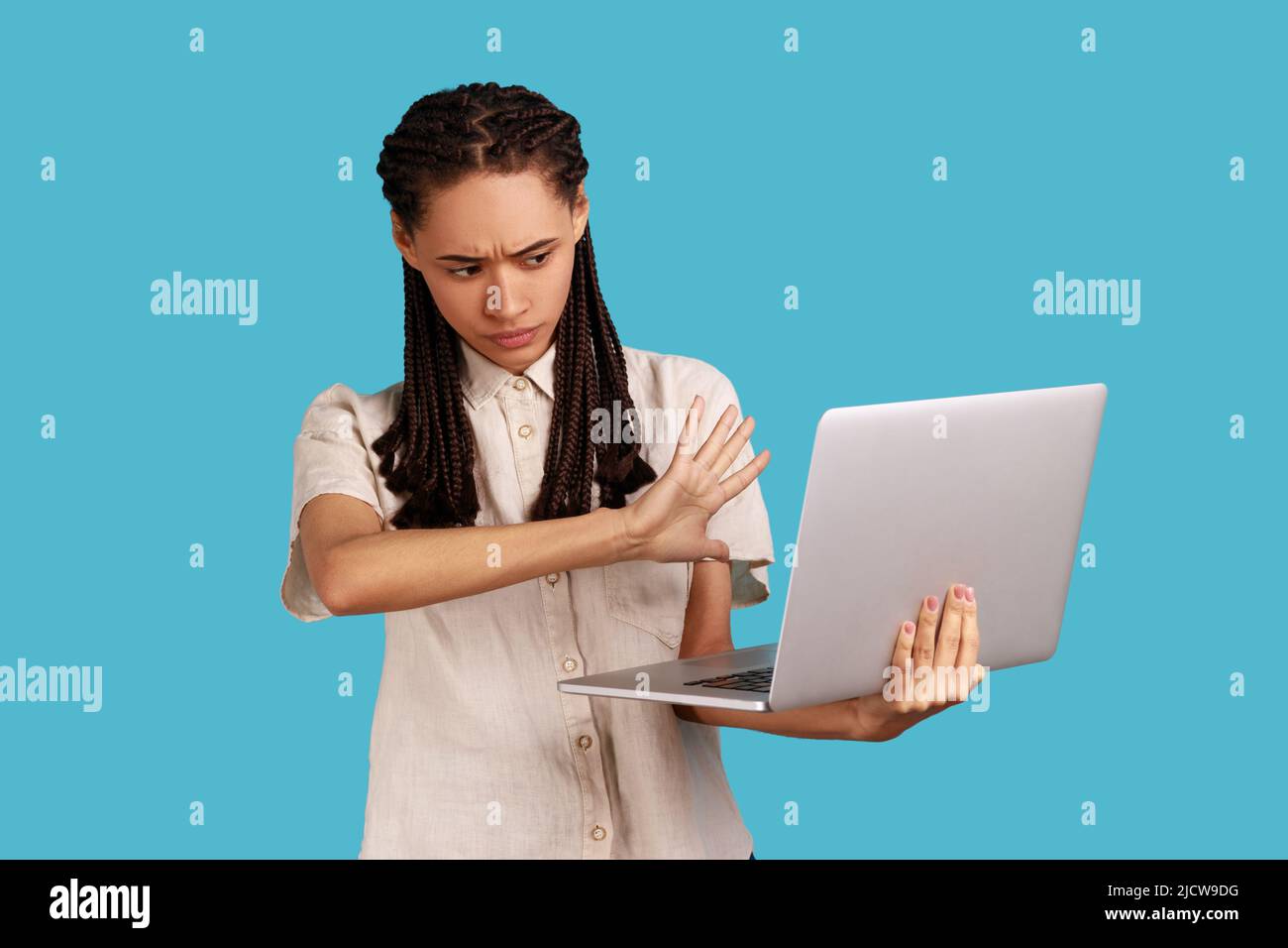 Portrait of woman with black dreadlocks showing stop gesture to laptop display, forbidden content, looking with disgust, wearing white shirt. Indoor studio shot isolated on blue background. Stock Photo