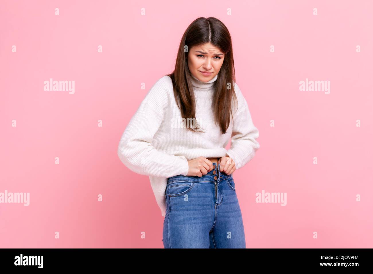 Portrait of unhappy sad young adult woman gaining weight, cant wearing her jeans, being overweight, wearing white casual style sweater. Indoor studio shot isolated on pink background. Stock Photo