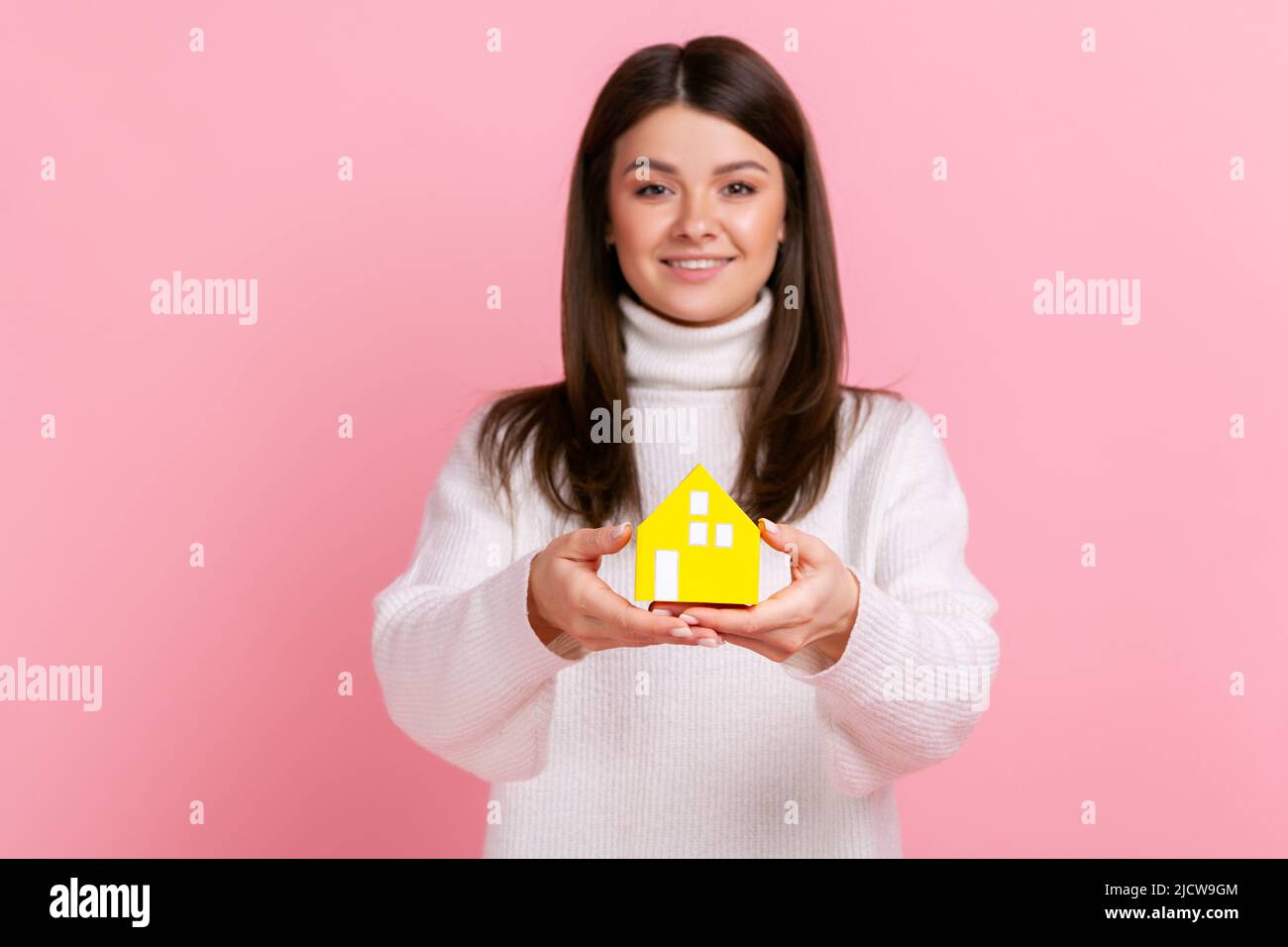 Beautiful woman with dark hair holding out paper small house, planning new home to buy, real estate, wearing white casual style sweater. Indoor studio shot isolated on pink background. Stock Photo
