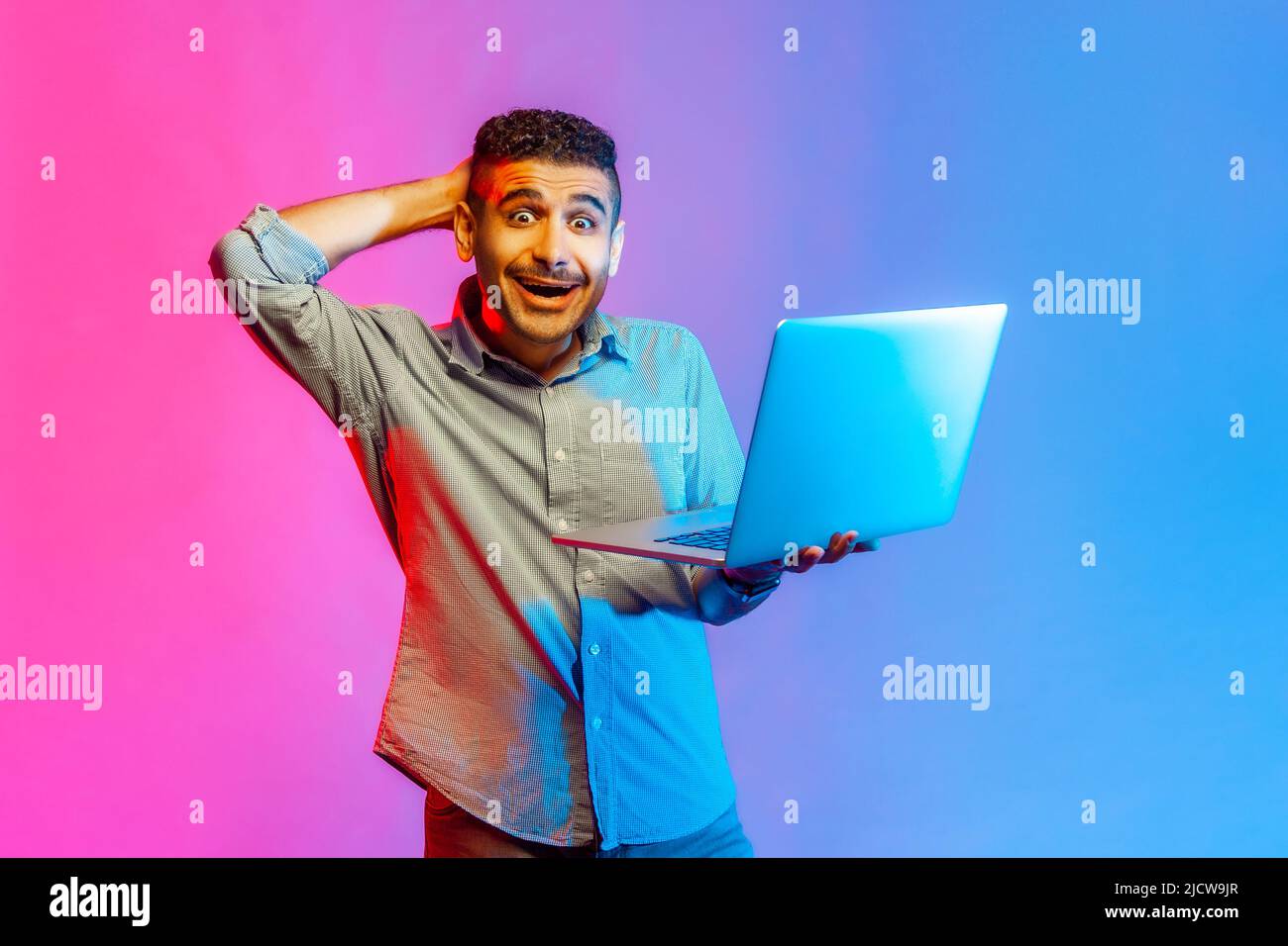 Portrait of man in shirt looking at camera with amazed expression, watching shocking content, surprised by news, raised arm. Indoor studio shot isolated on colorful neon light background. Stock Photo