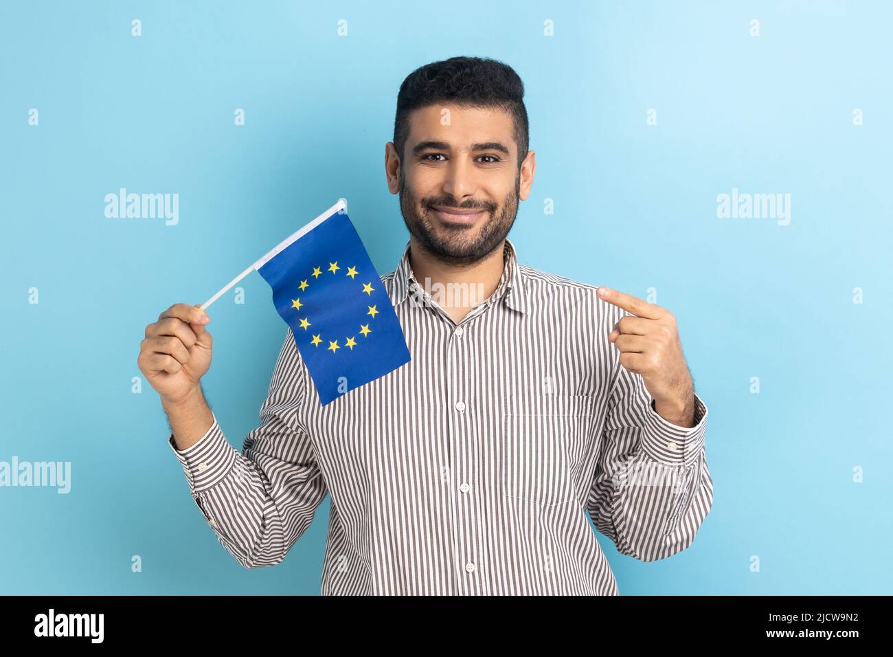Bearded businessman smiling broadly and pointing flag of European Union, symbol of Europe, EU association and community, wearing striped shirt. Indoor studio shot isolated on blue background. Stock Photo