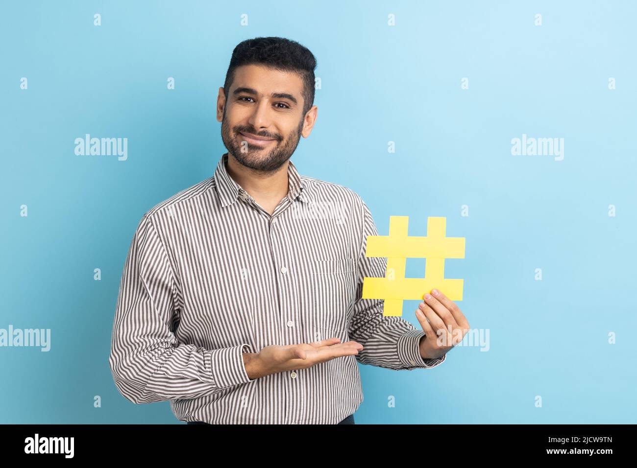 Portrait of smiling bearded businessman presenting yellow hashtag, tagging blog trends, viral topic in social network, wearing striped shirt. Indoor studio shot isolated on blue background. Stock Photo