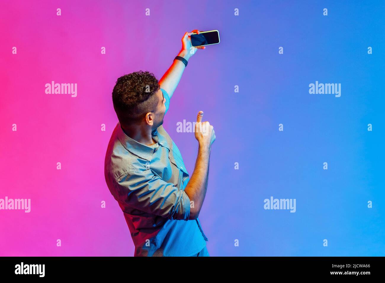 Back side of man in shirt taking selfie or broadcasting livestream, holding smart phone with blank screen, showing thumb up. Indoor studio shot isolated on colorful neon light background. Stock Photo