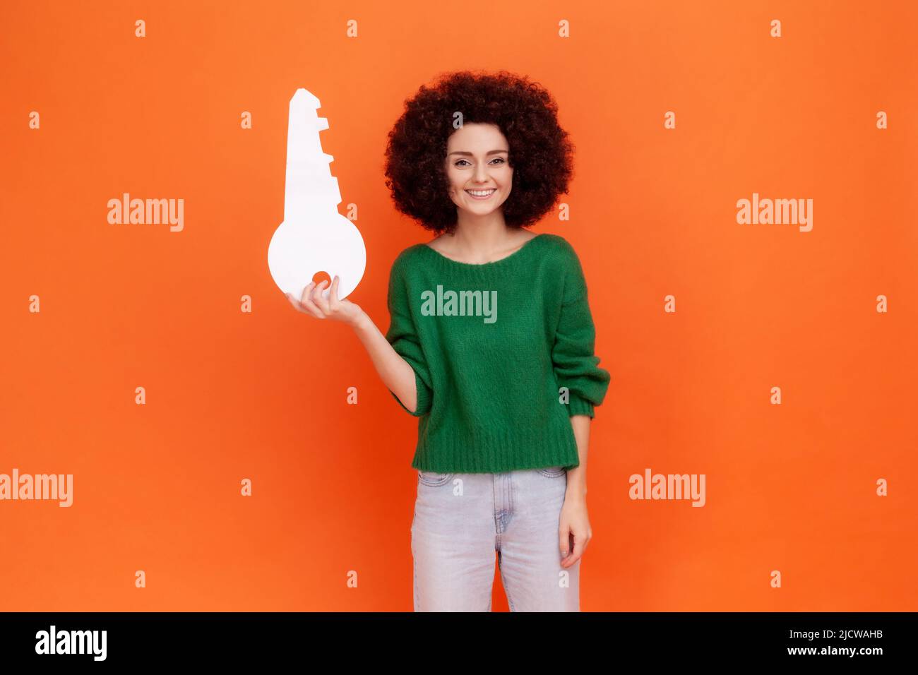 Portrait of smiling woman with Afro hairstyle wearing green casual style sweater holding paper key in hands, looking at camera, property. Indoor studio shot isolated on orange background. Stock Photo